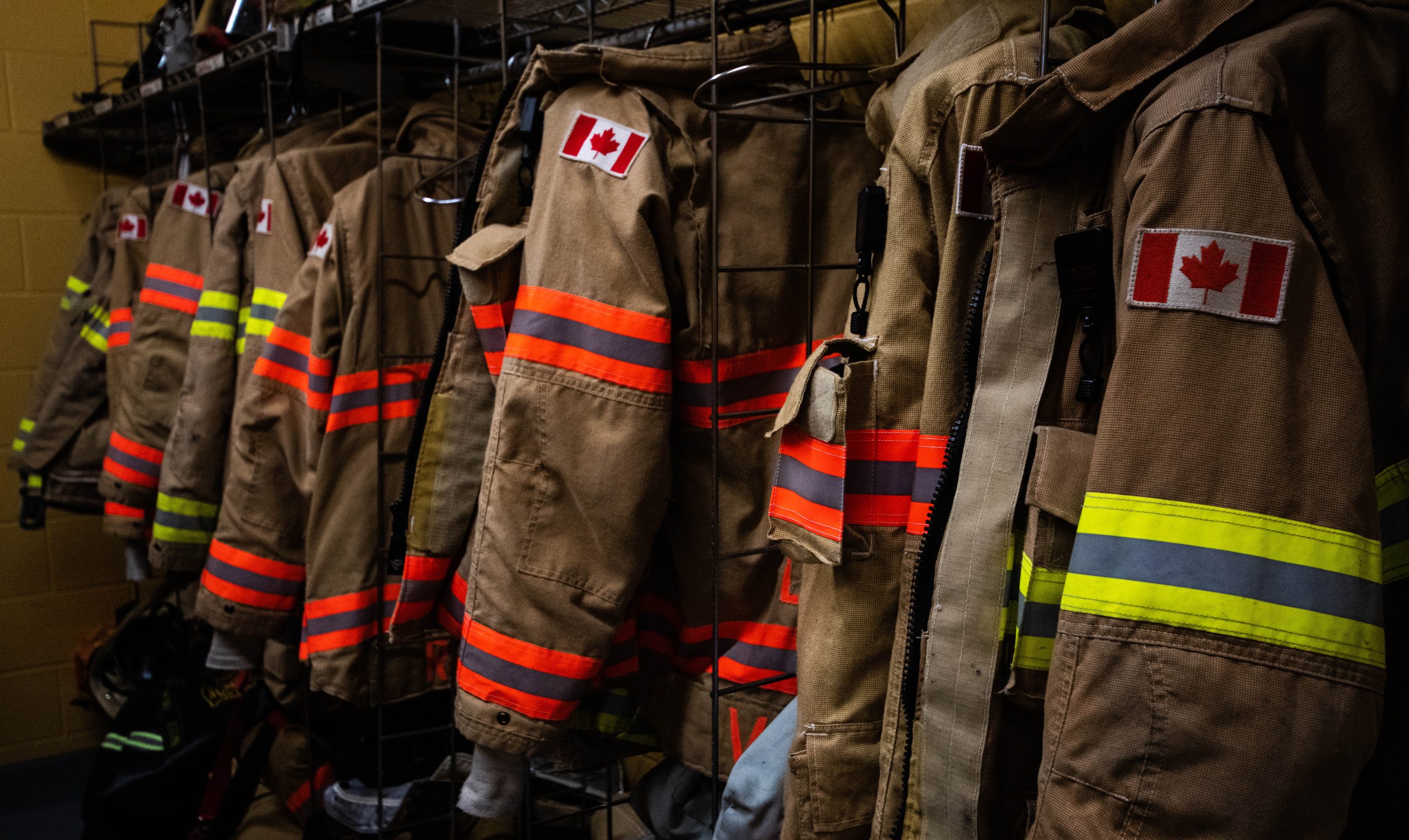 A rack of off-duty fire fighters’ bunker gear hanging on a rack waiting for the next call. Brown and beige with orange and yellow flourescent stripes and showing a Canadian flag.